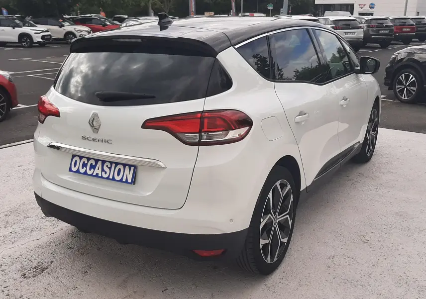 Vue 3/4 arrière droite d'une Renault Scénic IV blanc nacré avec toit noir, sur parking, badge et feux arrière visibles.
