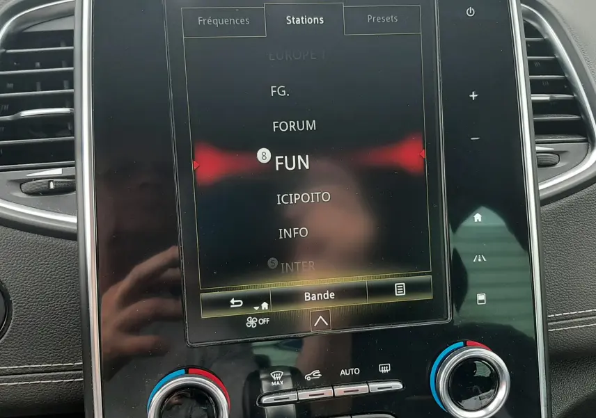 Vue rapprochée de l’écran tactile central du Renault Scénic IV blanc nacré, affichant les stations radio avec commandes climatisation en dessous.