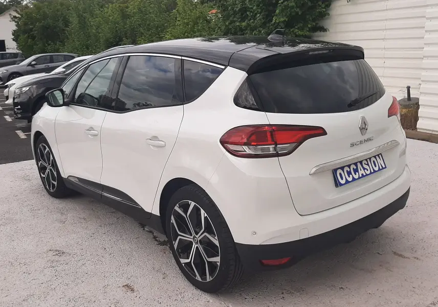 Vue 3/4 arrière droite d’un Renault Scénic IV blanc nacré avec toit noir et jantes alliage bicolores, stationné en extérieur.