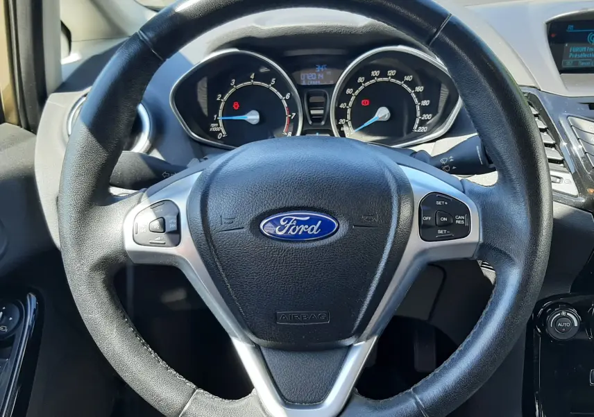 Vue intérieure centrée sur le volant noir de la Ford Fiesta marron métal, avec tableau de bord et commandes visibles.