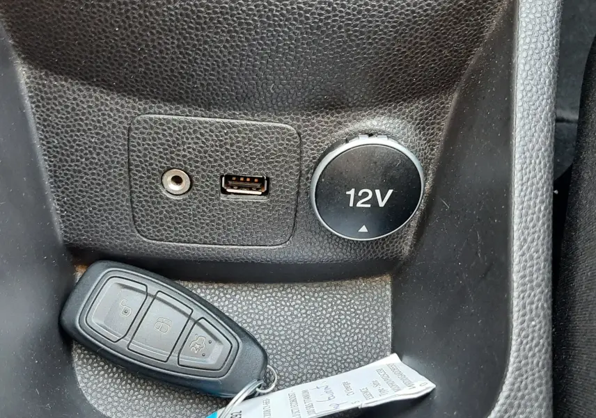 Gros plan sur la prise 12V, port USB et prise auxiliaire dans l’habitacle noir d’une Ford Fiesta marron métallisée.