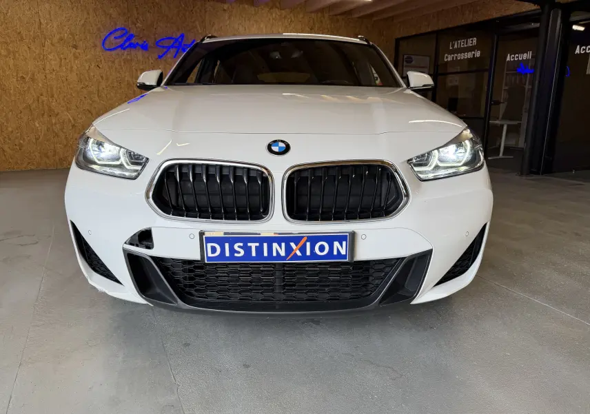 Vue frontale d'un BMW X2 blanc avec calandre noire et phares LED allumés dans un garage.