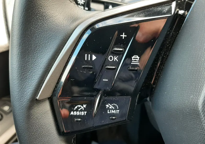 Gros plan sur les commandes multifonctions noires à gauche du volant cuir gris du Peugeot 5008 Hybrid 2025.