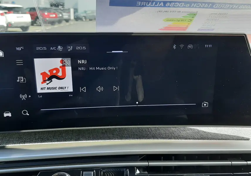 Vue intérieure du tableau de bord du Peugeot 5008 Hybrid 2025, écran tactile affichant la radio NRJ, avec bouton start/stop visible.