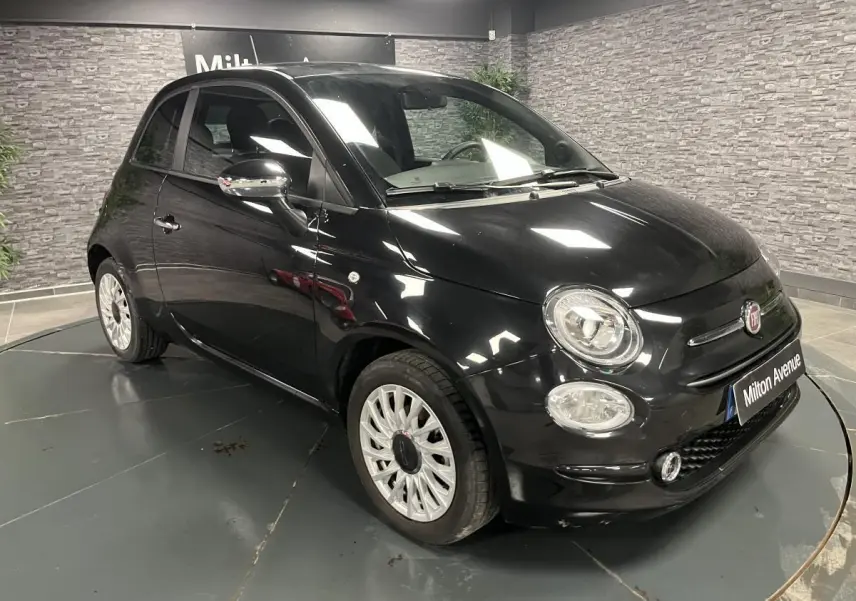 Fiat 500 noire vue en 3/4 avant droit, avec jantes alliage blanches et rétroviseurs chromés dans un showroom.