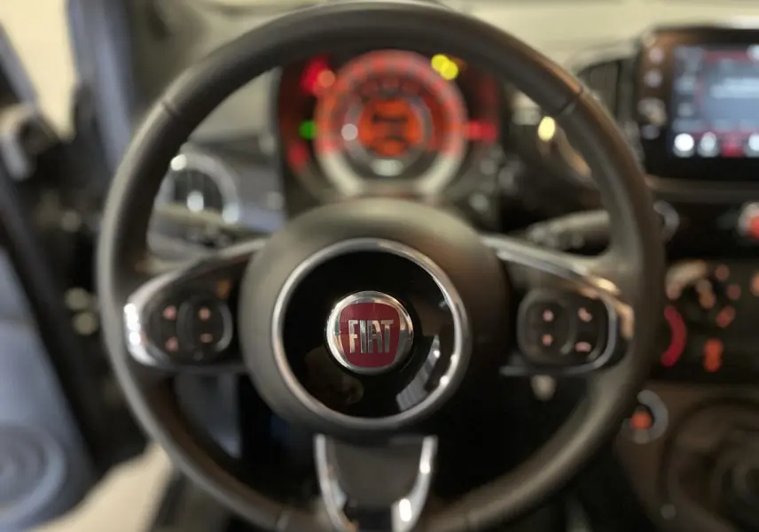 Volant noir de la Fiat 500 2023 avec logo central, commandes intégrées, tableau de bord flou en arrière-plan.