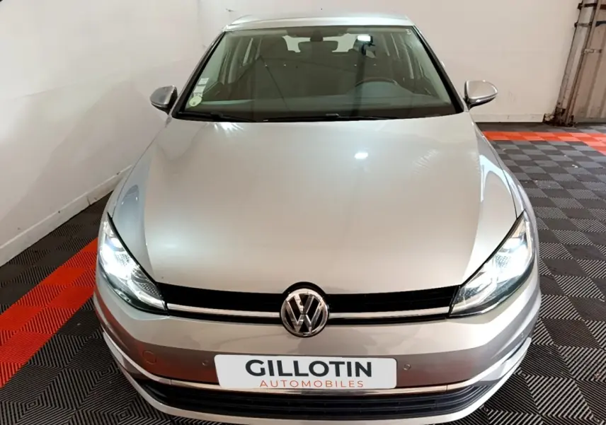 Vue avant d'une Volkswagen Golf gris clair avec phares allumés dans un garage au sol damier noir et rouge.