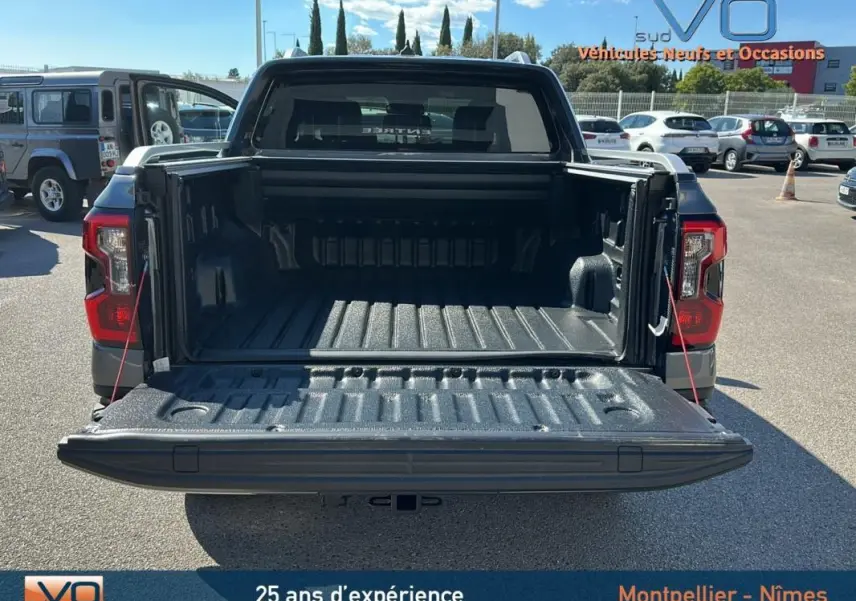 Vue arrière du Ford Ranger gris 2025 avec benne ouverte et hayon déployé sur parking ensoleillé.