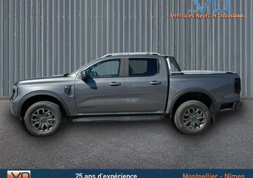 Profil côté gauche d'un Ford Ranger Wildtrak gris 2025 avec barres de toit et jantes alliage 17 pouces sur fond industriel.