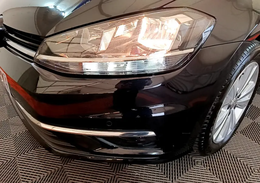 Gros plan sur l'avant droit d'une Volkswagen Golf noire 2018, mettant en valeur le phare allumé et la jante alu.