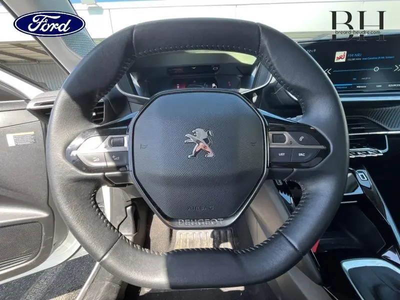 Vue rapprochée du volant noir de la Peugeot 208 blanc banquise, avec commandes intégrées et tableau de bord numérique.