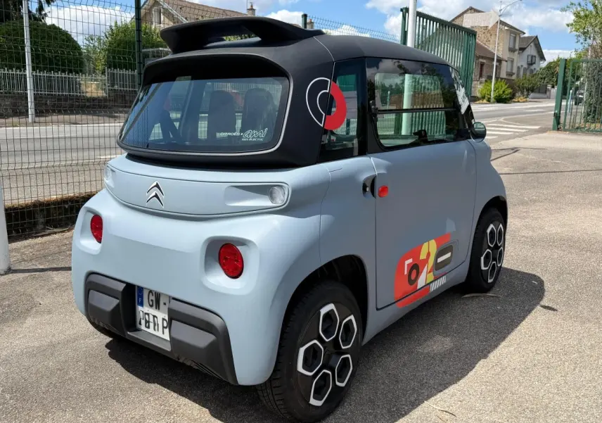 Vue 3/4 arrière droite d'une Citroën Ami MY AMI POP électrique gris clair avec toit noir et stickers colorés sur la porte.