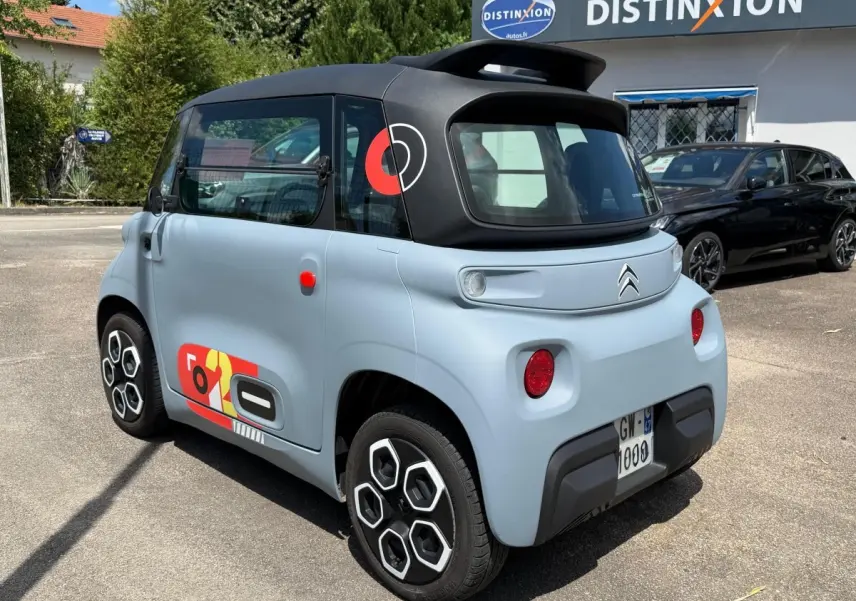 Vue 3/4 arrière droite de la Citroën Ami 2024 gris clair avec toit noir et stickers colorés sur porte et montant arrière.