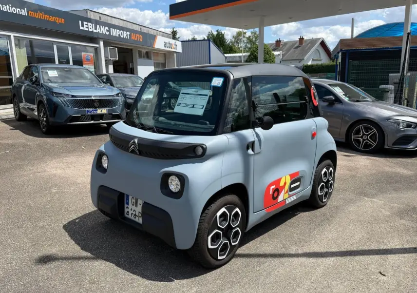 Vue 3/4 avant droite d'une Citroën Ami MY AMI POP électrique gris clair avec toit noir et stickers colorés sur porte.