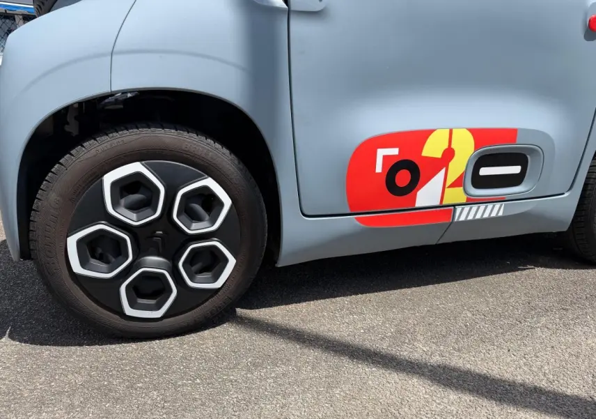 Gros plan sur la roue avant gauche et la porte côté conducteur d'une Citroën Ami MY AMI POP vert clair avec sticker rouge et jaune.