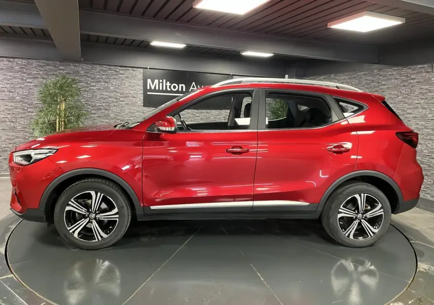 Vue de profil côté gauche du SUV MG ZS rouge métallisé avec jantes alliage noires et toit argenté en intérieur showroom.