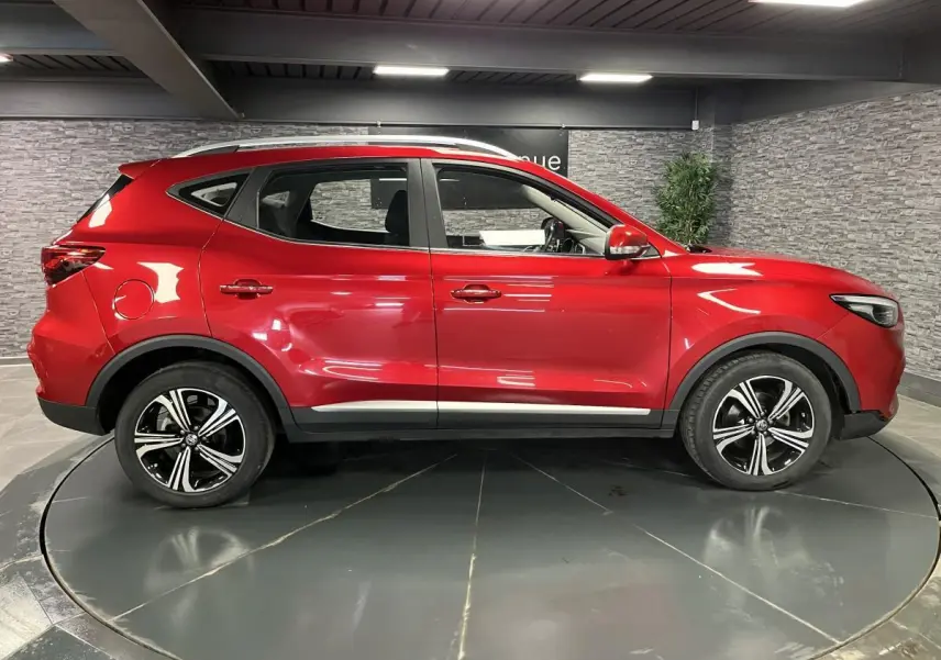 Vue latérale droite du SUV MG ZS rouge métallisé 2022 avec jantes alliage noires et toit argenté dans un showroom.