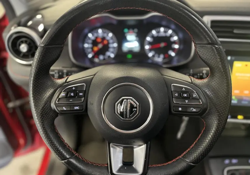 Gros plan sur le volant cuir noir avec surpiqûres rouges du MG ZS rouge, tableau de bord et commandes visibles en arrière-plan.