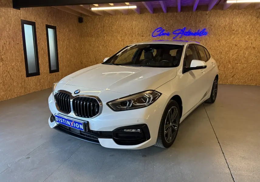 BMW Série 1 118i blanc en 3/4 avant droit, avec calandre chromée et jantes bi-ton dans un showroom.
