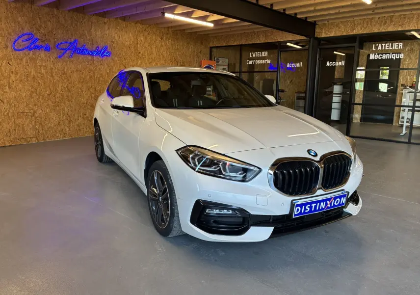 BMW Série 1 118i Edition Sport blanche vue en 3/4 avant droit dans un showroom avec calandre noire distinctive.