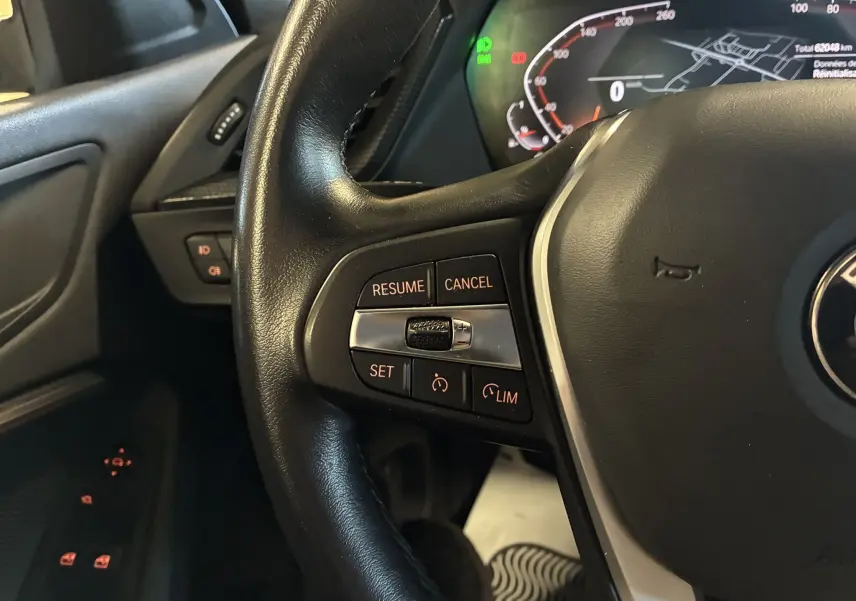 Gros plan sur le volant noir de la BMW Série 1 118i Edition Sport 2023, avec commandes de régulateur et tableau de bord numérique.