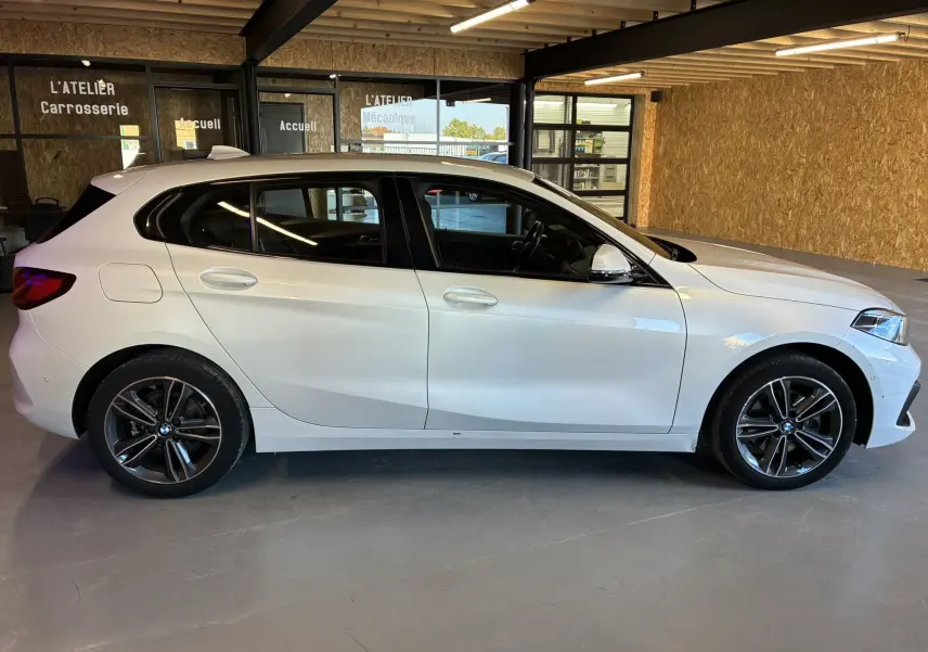 BMW Série 1 118i Edition Sport blanche vue de profil côté gauche dans un garage avec jantes bi-ton et vitres teintées.