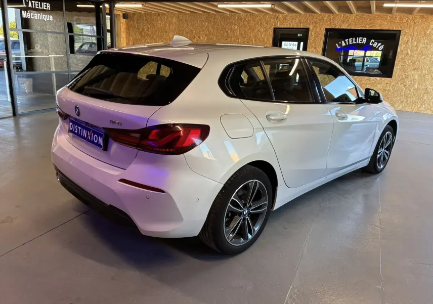 BMW Série 1 118i Edition Sport blanche vue en 3/4 arrière droit, avec jantes bi-ton et feux arrière LED allumés.