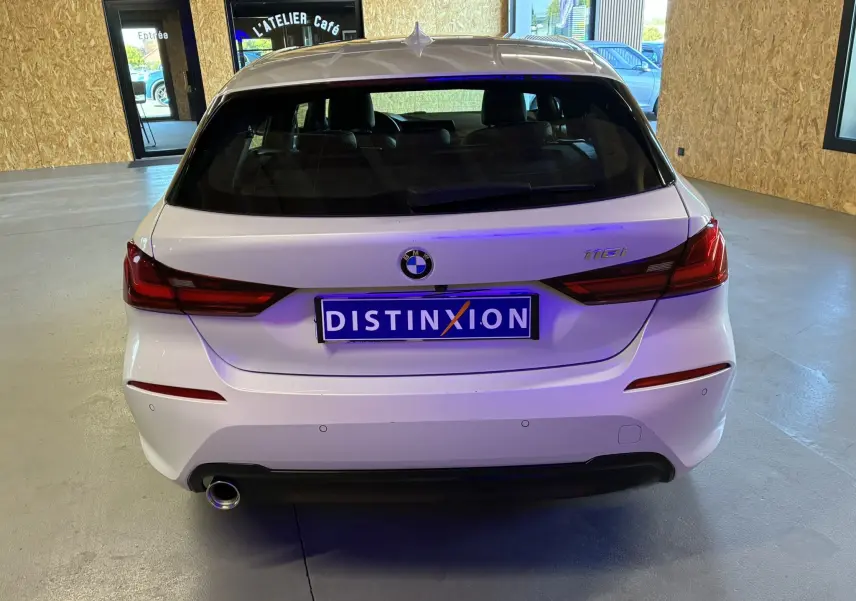 Vue arrière d'une BMW Série 1 118i blanche 2023 avec feux rouges et échappement visible en intérieur showroom.