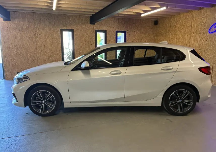 BMW Série 1 118i Edition Sport blanche vue de profil côté gauche dans un garage avec jantes bi-ton et vitres teintées.