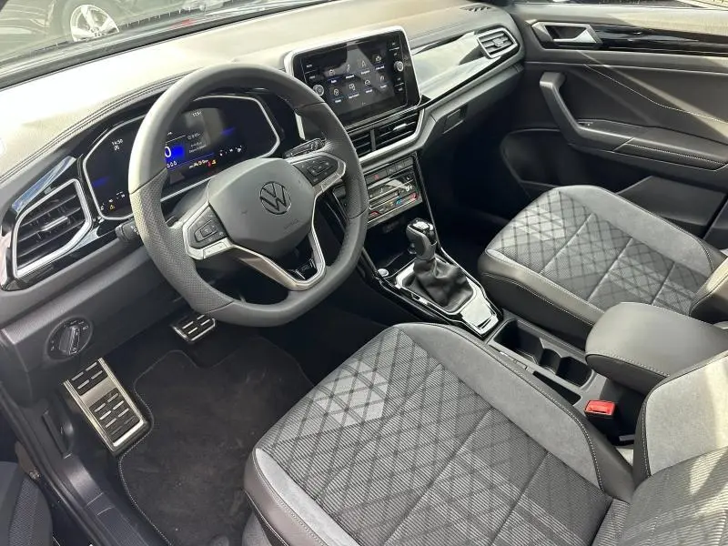 Vue intérieure côté conducteur du Volkswagen T-Roc 2025 avec volant multifonction, sièges tissu gris et console centrale moderne.