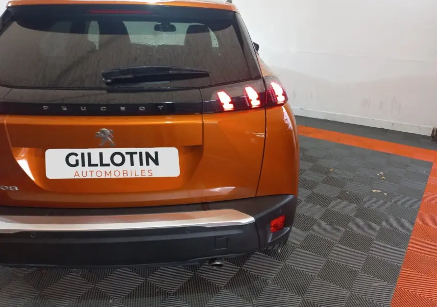 Vue arrière d'un Peugeot 2008 orange avec feux arrière allumés dans un garage au sol gris et orange.