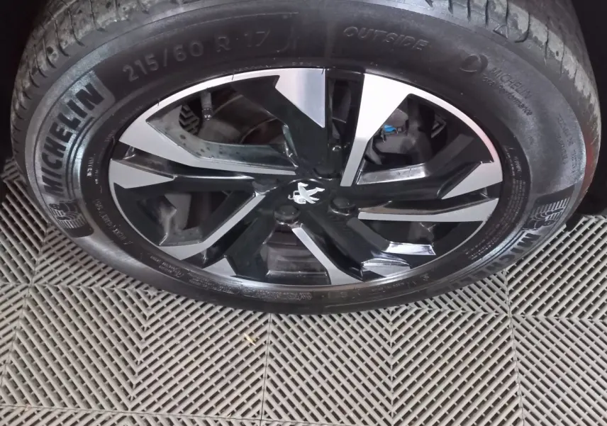 Gros plan sur la jante alliage noire et argentée avec logo Peugeot au centre, pneu Michelin visible sur sol carrelé.