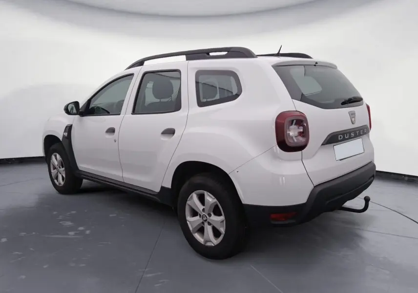 Vue 3/4 arrière droite d’un Dacia Duster blanc 2021 avec attelage et barres de toit noires visibles.