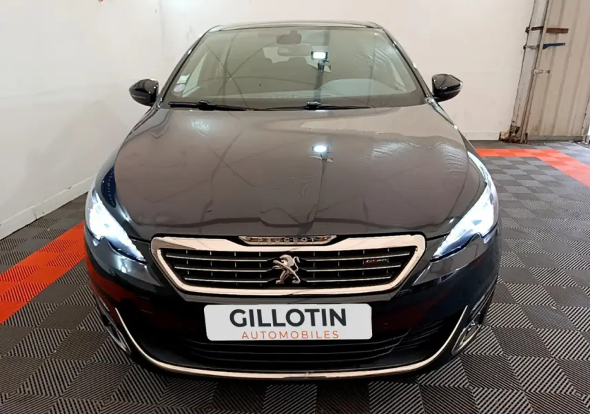 Vue frontale d'une Peugeot 308 gris foncé avec phares LED allumés et calandre chromée dans un garage.