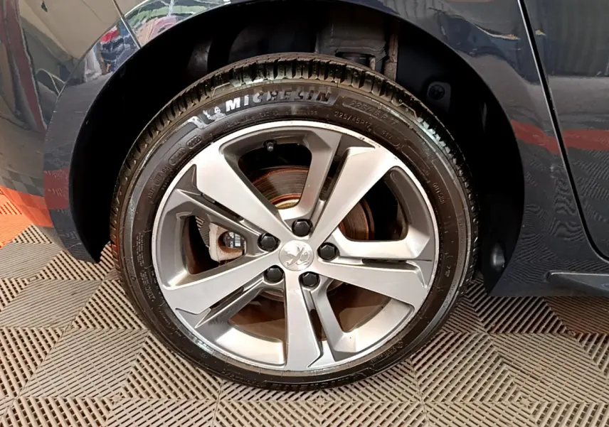 Gros plan sur la roue avant gauche avec jante alliage argentée et logo Peugeot sur une 308 gris foncé.