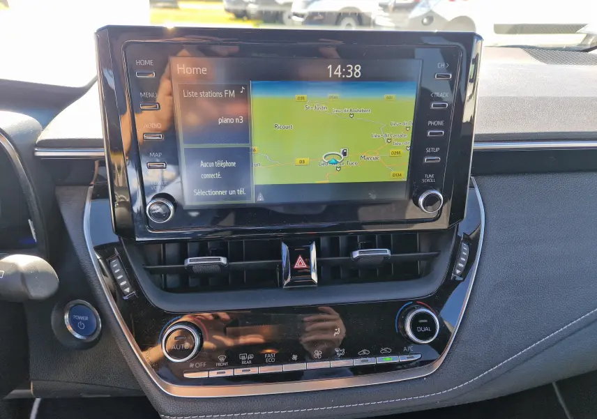 Vue rapprochée de l’écran tactile central et de la console de la Toyota Corolla Hybride 122h gris clair, avec commandes climatisation et bouton démarrage.