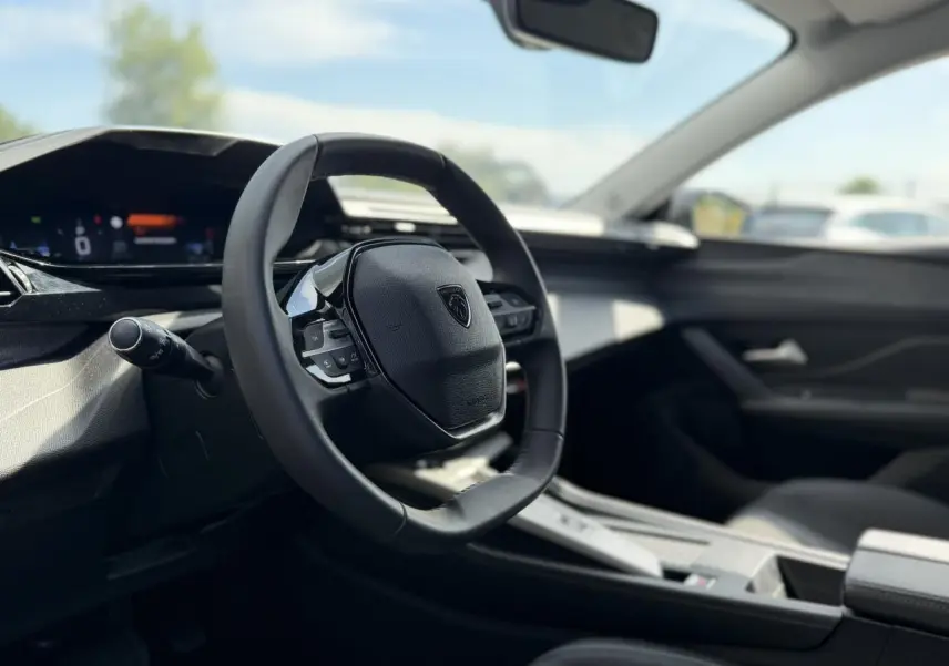Intérieur de la Peugeot 308 SW 2025 vu côté conducteur, volant cuir noir avec logo Peugeot au centre.