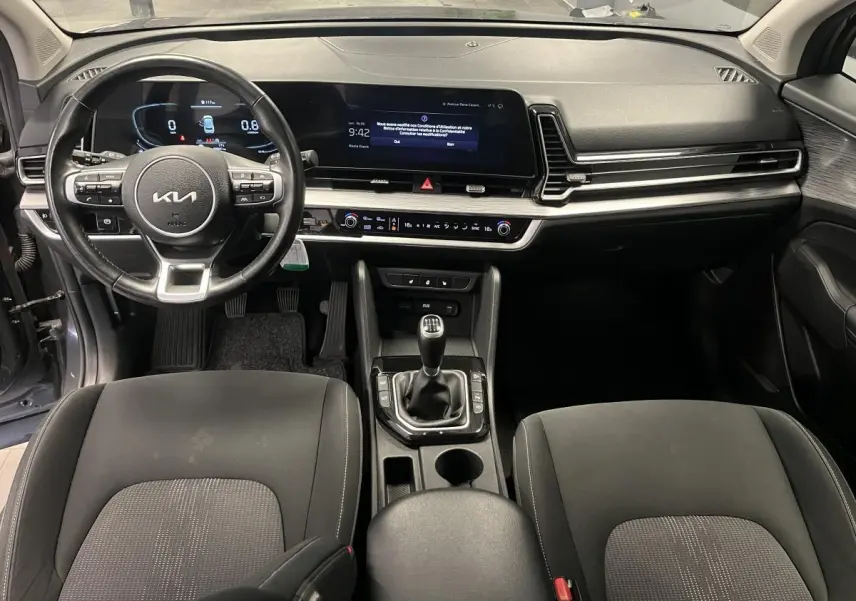 Vue intérieure avant du Kia Sportage 2022 gris, volant cuir, tableau de bord numérique et boîte manuelle visible.