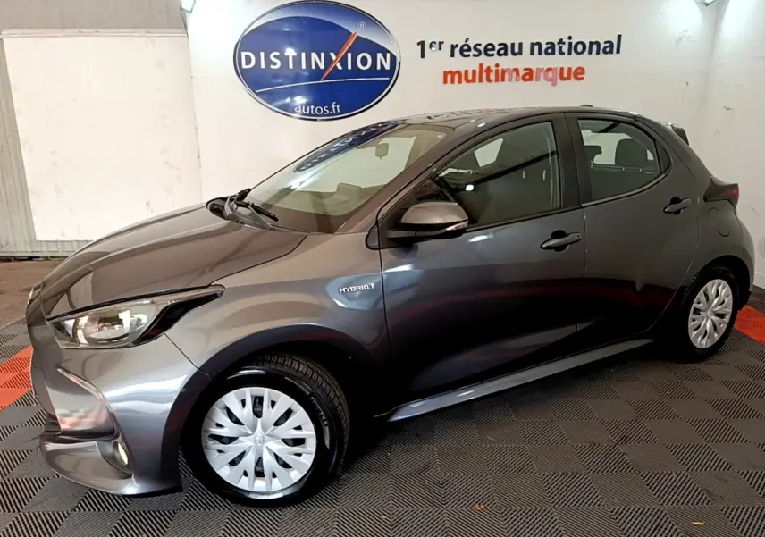 Toyota Yaris hybride gris clair vue de profil côté gauche, avec logo Hybrid et feux avant allumés.