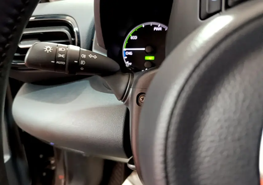 Gros plan sur le compteur hybride et le commodo d'éclairage à gauche du volant cuir d'une Toyota Yaris gris clair 2021.