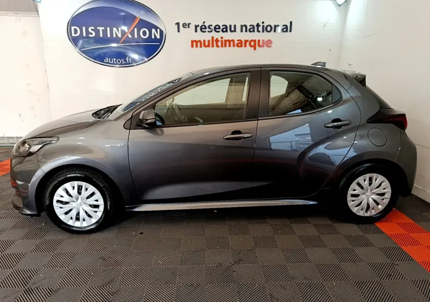 Profil côté gauche d'une Toyota Yaris hybride gris clair 2021, avec jantes à enjoliveurs blancs et logo hybride visible.