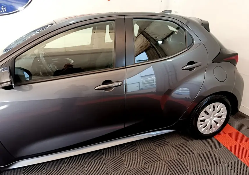Profil côté gauche d'une Toyota Yaris hybride gris clair, avec jantes à enjoliveurs et poignées de porte assorties.