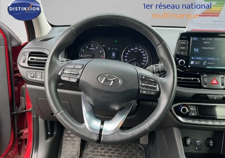 Vue intérieure centrée sur le volant cuir d’une Hyundai i30 SW rouge, avec tableau de bord et écran multimédia visibles.