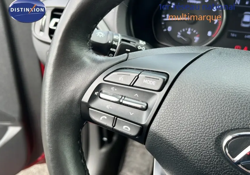 Gros plan sur le volant côté gauche de la Hyundai i30 SW 2023 en rouge Sunset Red, avec commandes audio intégrées.