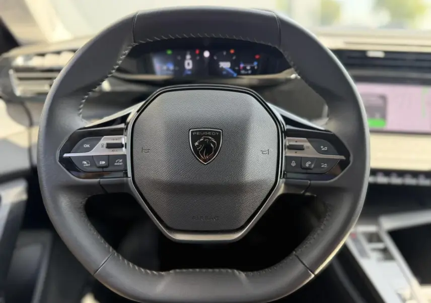 Vue rapprochée du volant cuir noir de la Peugeot 308 SW 2025 avec commandes intégrées et tableau de bord digital flou.