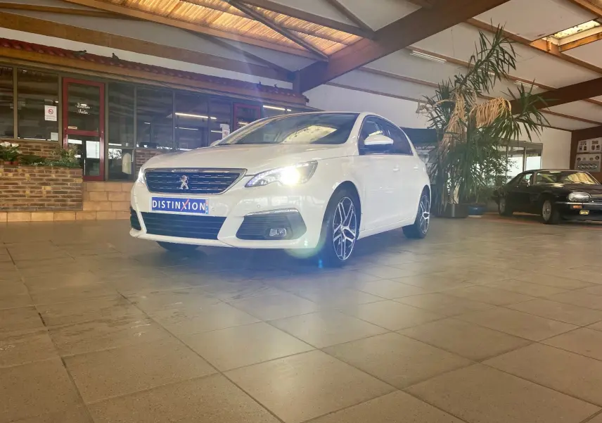 Peugeot 308 blanche vue en 3/4 avant droit dans un showroom, phare avant droit allumé et jantes alliage visibles.