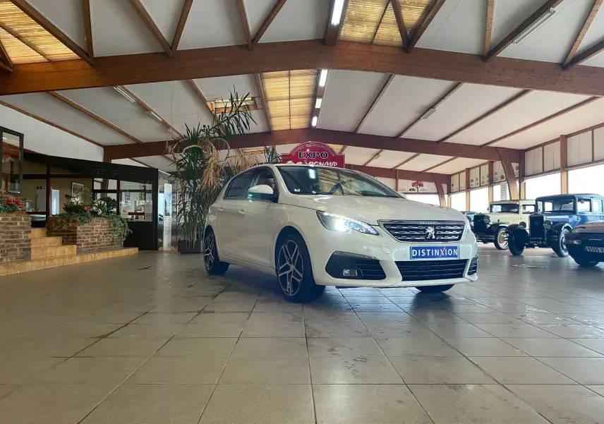 Peugeot 308 blanche vue en 3/4 avant droit dans un showroom avec des voitures anciennes en arrière-plan.