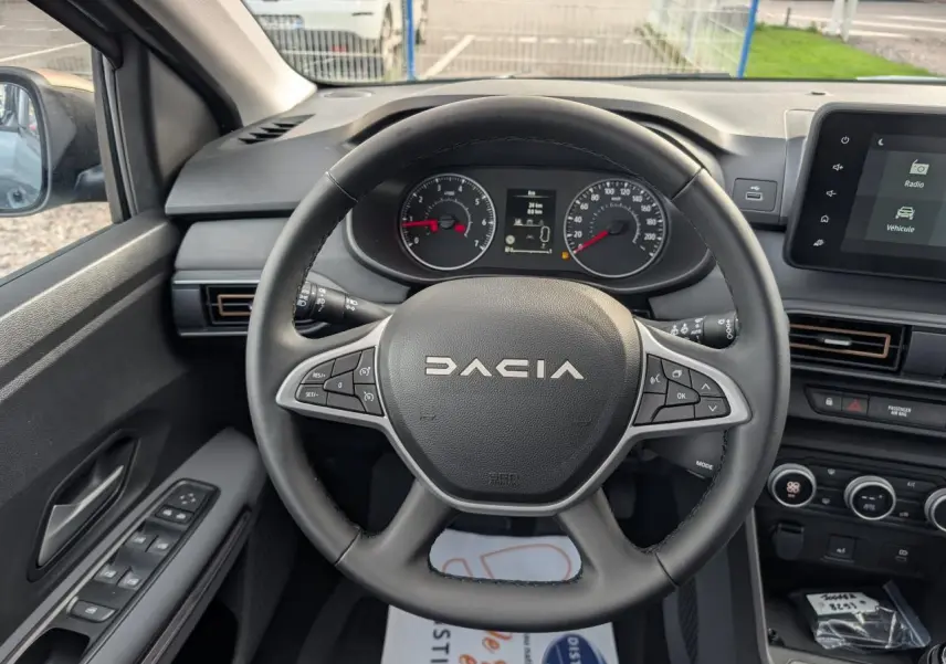 Vue intérieure centrée sur le volant DACIA noir avec tableau de bord et écran tactile à droite, portière côté gauche visible.