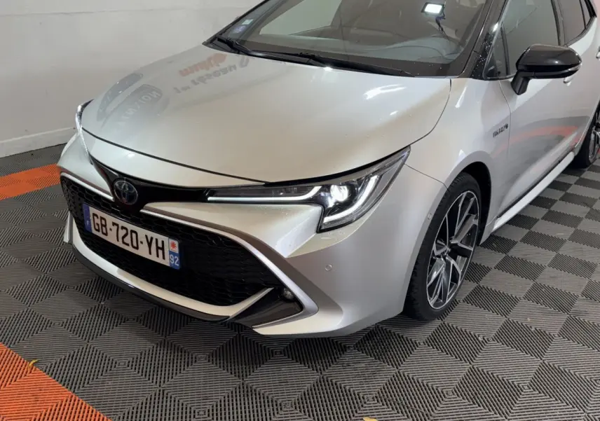 Vue 3/4 avant droit d'une Toyota Corolla hybride gris clair avec phares LED allumés et jantes alliage distinctives.