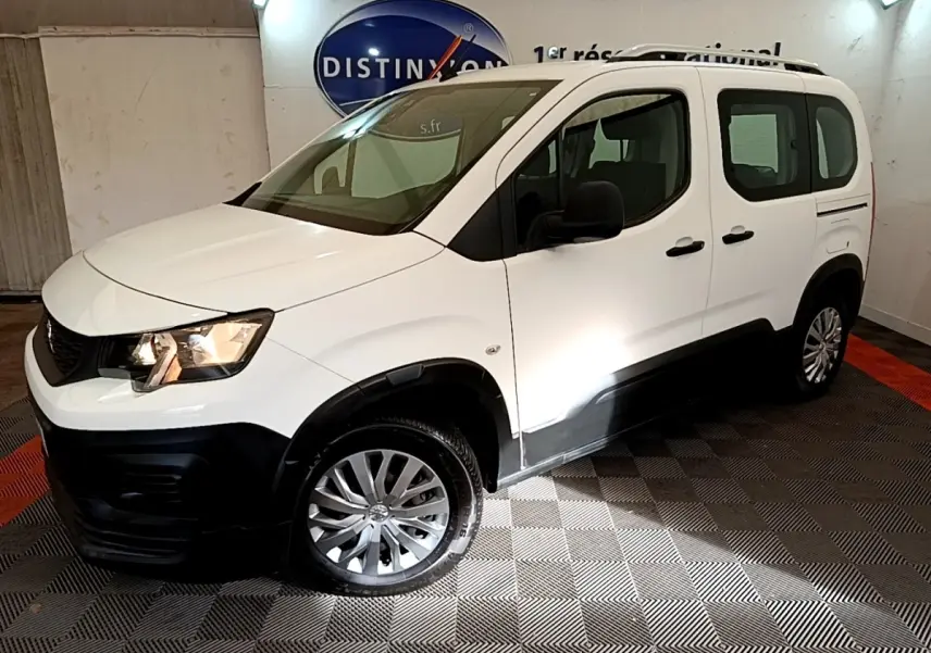 Vue 3/4 avant gauche d’un Peugeot Rifter blanc avec protections noires et jantes alliage, en intérieur showroom.